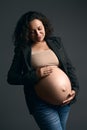 Authentic pregnant woman in stylish grey blazer and blue jeans, smiling, stroking her bare belly, on studio background