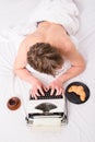 Author guy tousled hair busy write chapter deadline coming top view. Man with typewriter and coffee lay bed. Man