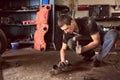 Strong auto mechanic in dirty workwear squatting working on car detail on floor with hammer helping at auto repair shop