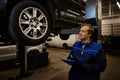 Auto mechanic, technician in uniform making the checklist on clipboard for repairing the car lifted on a hoist in the repair shop Royalty Free Stock Photo