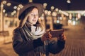 Autumn evening. Young woman in hat and scarf stands on city street, uses smartphone. Hipster girl uses digital gadget.