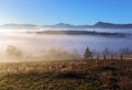 Autumn landscape of high mountains, orange coloured trees, fog. Sun rays enlighten the lawn with dry grass. Blue sky. Royalty Free Stock Photo