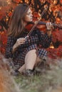 Autumn portrait of beautiful woman sitting on the ground with a violin under chin on a background of red foliage, girl engaged in