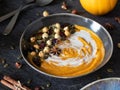 Autumn pumpkin soup in a bowl with coconut cream, pumpkin seeds, hazelnuts, and dry cranberries on a grey table with spoons and ci