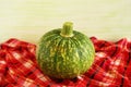 Still life with green pumpkin in a center on a checkered towel on a light green wooden background.