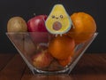 An avocado cut in half drawn on a sheet of paper is placed against a glass container with oranges, kiwis, apples and walnuts