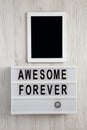 `Awesome forever` word on modern board, tablet with blank screen on a white wooden surface, top view. From above, flat lay,