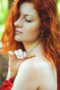 Awesome girl with freckles and red hair posing with a lowered red dress and bare shoulders on wild nature.