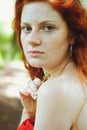 Awesome girl with freckles and red hair posing with a lowered red dress and bare shoulders on wild nature.