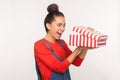 Awesome holiday gift! Portrait of surprised nosy pretty girl with hair bun in denim overalls looking into present box