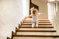 Baby blonde girl in white t-shirt at bottom of stairs indoors looking at camera and smiling