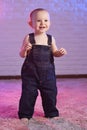 baby boy in denim overalls smiling while standing barefoot on the carpet against the background of a white brick wall Royalty Free Stock Photo