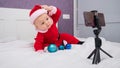 A baby boy dressed in a Santa suit sits on the bed and looks at the sick phone. Christmas greetings by video call Royalty Free Stock Photo