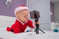 A baby boy dressed in a Santa suit sits on the bed and looks at the sick phone. Christmas greetings by video call Royalty Free Stock Photo