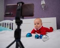 A baby boy dressed in a Santa suit sits on the bed and looks at the sick phone. Christmas greetings by video call Royalty Free Stock Photo