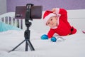 A baby boy dressed in a Santa suit sits on the bed and looks at the sick phone. Christmas greetings by video call Royalty Free Stock Photo