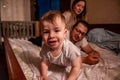 Baby boy lies on stomach on bed, smiling at camera. Family efocused from behind. Care of children Royalty Free Stock Photo