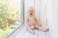 Baby boy 8 months old sitting in diapers on the window sill and looking into the distance, a place for text Royalty Free Stock Photo