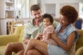 Baby Daughter Sitting With Parents Loving On Sofa At Home With Grandparents In Background Royalty Free Stock Photo