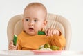 A baby on a feeding chair with two hands eats a fresh green cucumber. On a white background Royalty Free Stock Photo