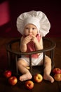 Baby in a chef& x27;s hat sits in a chair with a bunch of apples and eats something tasty