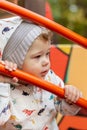 Baby girl boy in a hoodie and hat looks sideways through a metal fence. Caucasian child on a walk holds on to the railing Royalty Free Stock Photo