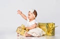 Baby girl in dress and headband, barefoot. She pulling hand, holding golden gift box, sitting on floor isolated on white. Close up Royalty Free Stock Photo