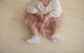 Baby girl in a dusty pink tulle skirt, white lace socks, with puffy arms, sits on a light-colored wooden floor