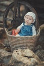 Baby girl with ocean deep blue eyes and pinky cheek wearing white shirt and jeans dress and handmade craft tiny hat sitting in bas