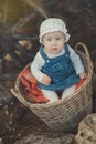 Baby girl with ocean deep blue eyes and pinky cheek wearing white shirt and jeans dress and handmade craft tiny hat sitting in bas