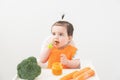 Baby girl in orange bib sitting in a Childs chair eating vegetable puree on white background