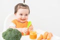Baby girl in orange bib sitting in a Childs chair eating vegetable puree on white background