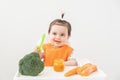 Baby girl in orange bib sitting in a Childs chair eating vegetable puree on white background