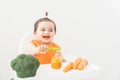 Baby girl in orange bib sitting in a Childs chair eating vegetable puree on white background