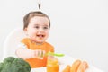 Baby girl in orange bib sitting in a Childs chair eating vegetable puree on white background