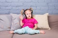 Baby girl sitting on sofa with pillows and listening to music in big headphones put on head. Royalty Free Stock Photo