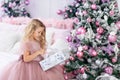 Baby girl unpacks a Christmas gift at the Christmas tree in a beautiful dress of pink color, the concept of new year and Christmas