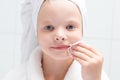 Baby girl, in a white robe and towel on her head, cleans her face with a round cotton swab, close-up, against the wall in the Royalty Free Stock Photo