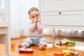 The baby hides his smile with his hands while sitting on the floor and playing with toys