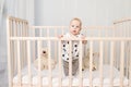 A baby of 8 months stands in a crib with toys in pajamas in a bright children`s room after sleeping and looks at the camera, a Royalty Free Stock Photo