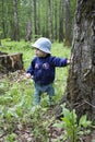Baby 8-9 months takes the first steps in nature. A girl in the woods looks around holding onto a tree trunk. Girl in jeans and a