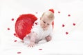 A baby with plump cheeks in white clothes with red cupid wings, plays with a heart, isolated on a white soft blanket with hearts. Royalty Free Stock Photo