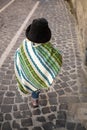 Baby in poncho and felt hat walking ahead along the street with cobblestone pavement. View from behind, no face. Baby