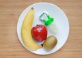 Baby`s nibbler and fresh ripe fruits apple, banana and kiwi on white plate against wood board background Royalty Free Stock Photo