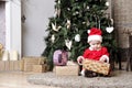 Baby in Santa costume sit near decorating Christmas tree and try unwrap present box
