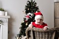 Baby in Santa costume stand on chair near Christmas tree