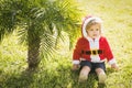 Baby santa, cute boy or child in red new year coat with white fur celebrates christmas near green palm tree on grass