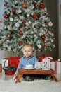 Baby is sitting at small tea table made against the background of beautiful Christmas tree