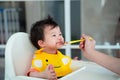Baby smiling while eating with Mother hand holding small spoon Feeding Baby,toddler is messy wearing yellow bib apron and sitting Royalty Free Stock Photo