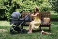 Baby Strollers for newborn. Young mother with newborn baby in stroller sitting on bench in the park. Outdoor portrait of